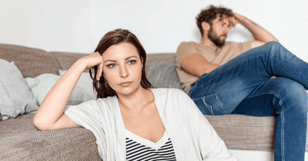 A woman sits on the floor looking frustrated while a man sits distant on the couch behind her, reflecting the reality that you’ll never change a narcissistic partner.