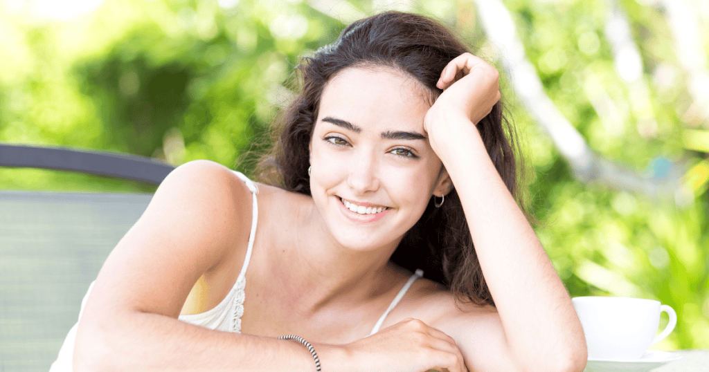 A young woman with long dark hair smiles while leaning on her arm outdoors beside a white cup, highlighting the idea that your radiance is the key to your freedom.