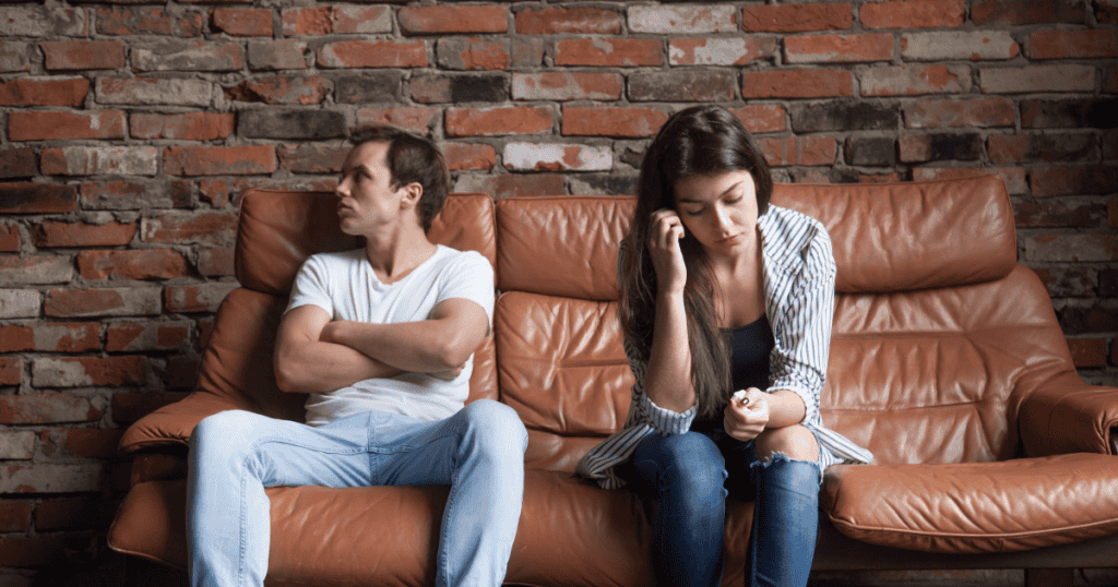 A couple sits in cold silence on opposite ends of a couch, capturing the manipulative tension narcissists create after a breakup.