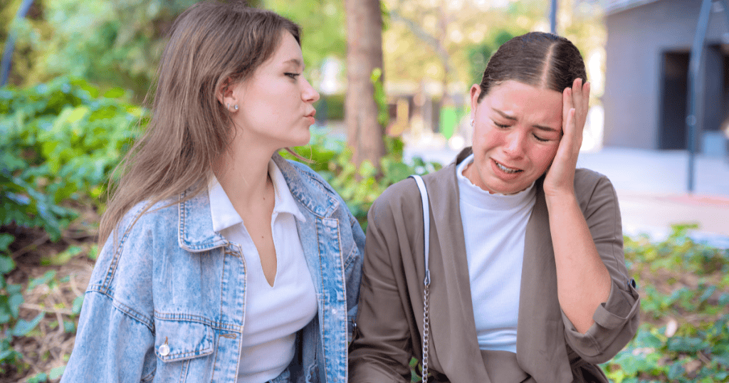 A woman sits outdoors looking upset with her hand on her forehead, while another woman beside her shakes her head quietly, observing her reaction.