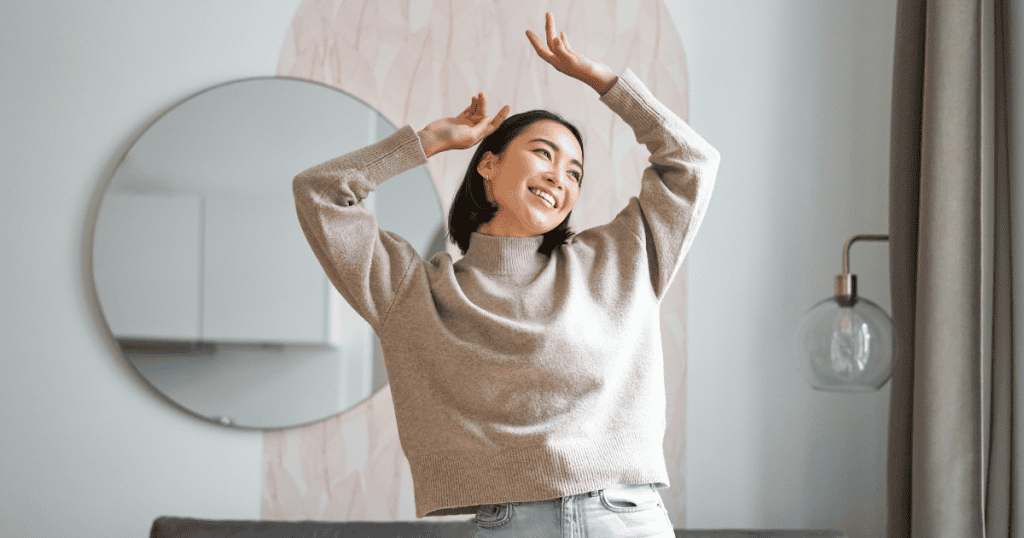 A woman dances joyfully alone in her living room with her arms raised, finding complete fulfillment within herself without needing external validation.