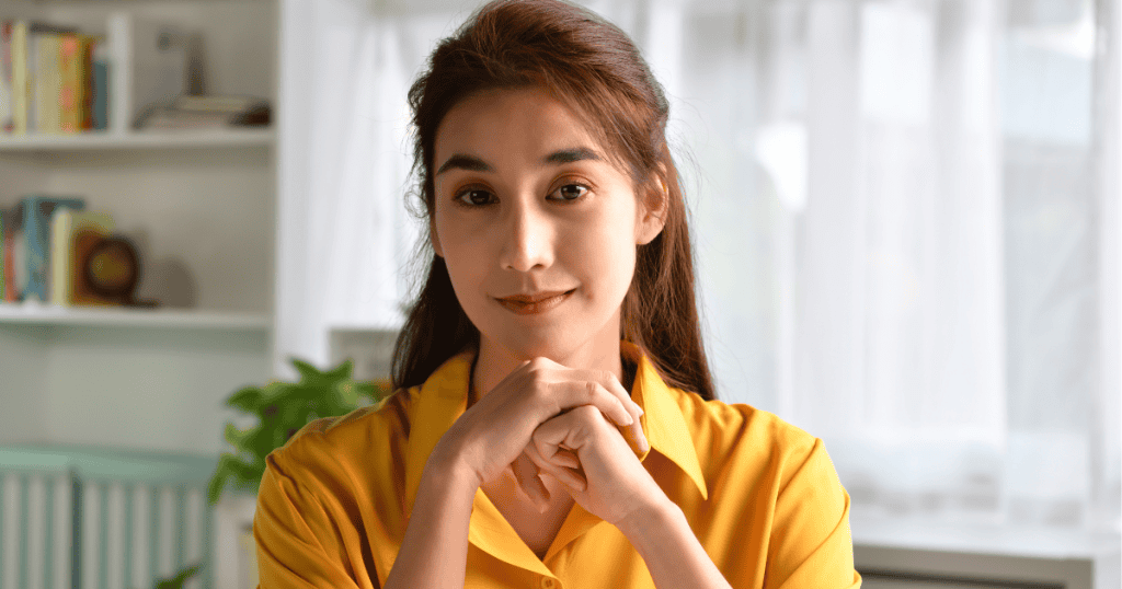 A woman in a yellow shirt rests her chin on her clasped hands with a gentle smile, projecting a calm and patient willingness to overlook flaws.