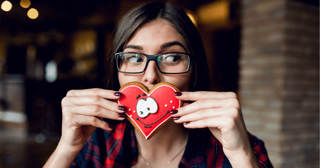 A woman playfully holds a decorated heart cookie over her mouth, keeping a sharp eye out for signs of genuine sweetness in her life.