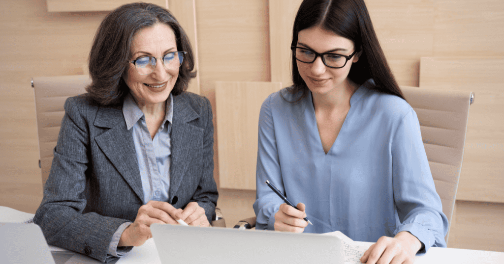 An older woman smiles while consulting with a younger professional at a desk, projecting an outward appearance of engagement that feels more like a calculated performance than a reflection of genuine worry.