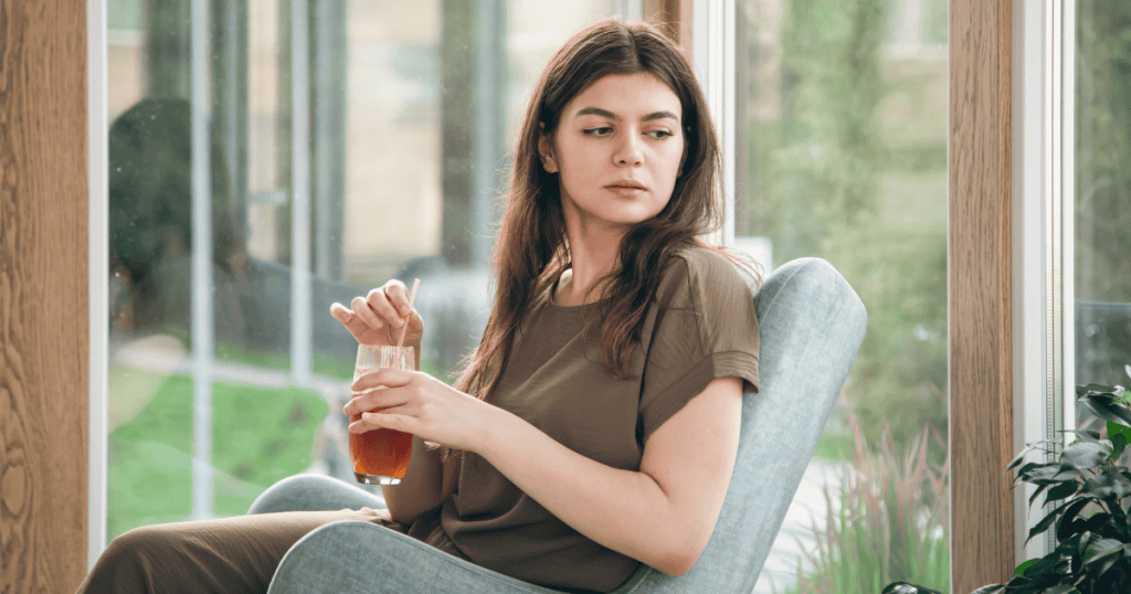 A woman sits in a chair and looks thoughtfully over her shoulder toward a window, signaling a shift in perspective and internal vigilance.