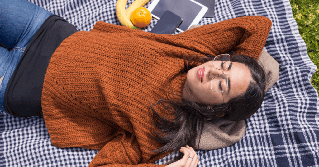 A woman lies peacefully on a picnic blanket with her eyes closed, embracing a sense of wholeness that doesn't need to be lessened for others.