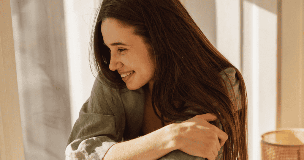 A woman smiles softly while hugging her knees in the sunlight, embracing the internal peace that comes with a profound realization.