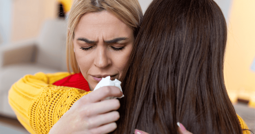 A tearful woman receives a hug while holding a tissue, her pained expression suggesting the comfort offered feels empty despite the physical closeness.