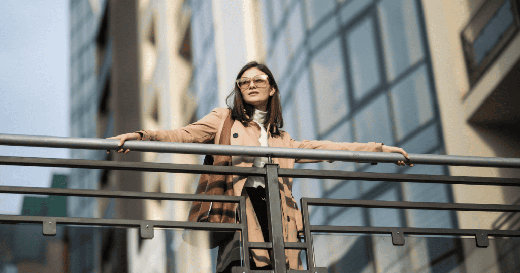 A woman in a trench coat stands confidently behind a metal railing against a backdrop of modern glass buildings, embodying the quiet strength of a self-reliant life.