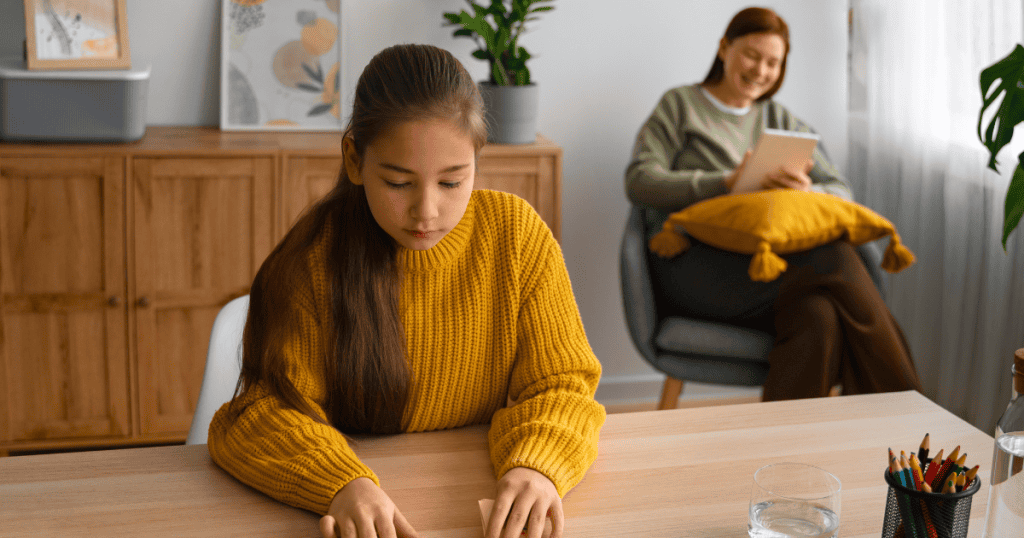 A girl focuses intently on her desk work while a woman watches from the background, reflecting a dynamic where a child’s efforts serve another's agenda.