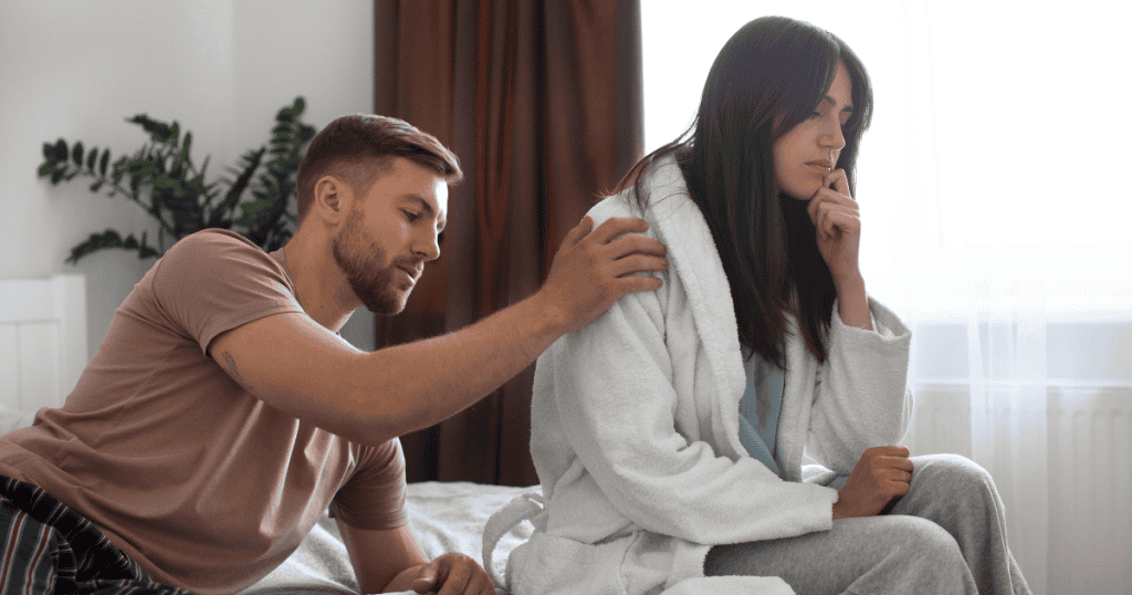 A man reaches out to touch a woman’s shoulder while she sits turned away in a white robe, capturing a moment of quiet tension and growing internal doubt.