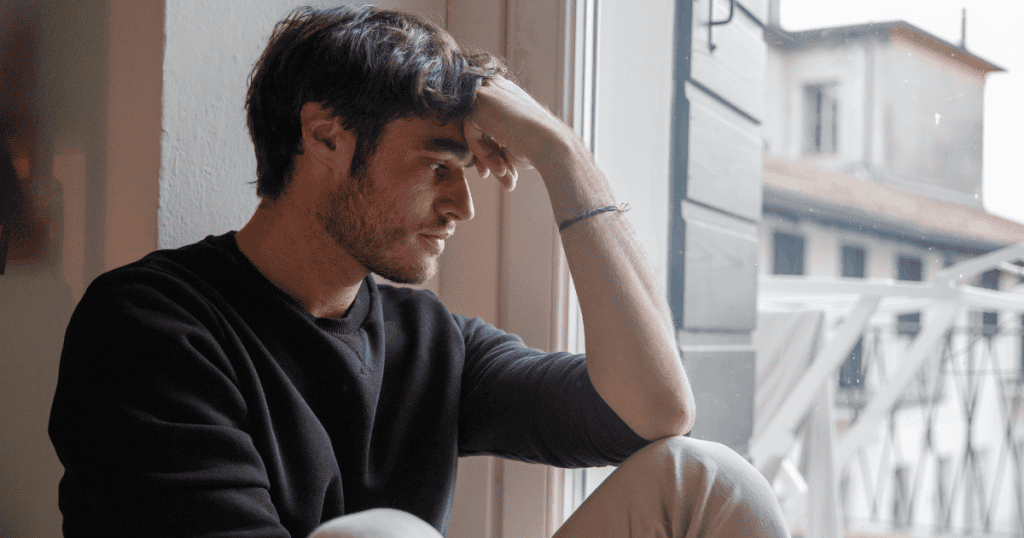 A young man sits by a window with his hand pressed to his forehead in deep thought, reflecting the heavy realization of a presence that is no longer there.