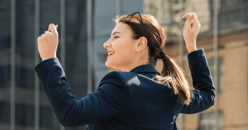 A young woman in a blazer smiles brightly with her arms raised in celebration against a city backdrop, showcasing the profound independence that flourishes once an external source of validation is removed.