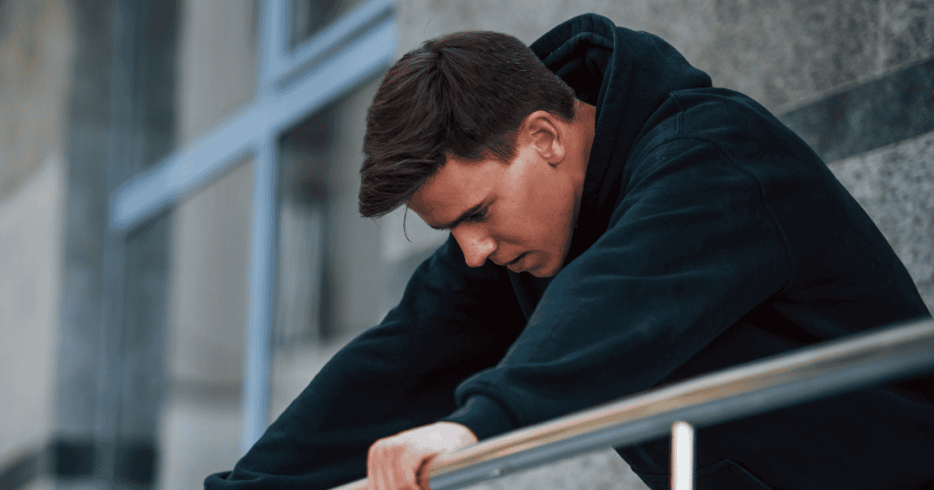 A young man in a dark hoodie leans heavily against a metal railing with a defeated stare, representing the visible cracks in a facade when external validation is suddenly withdrawn.