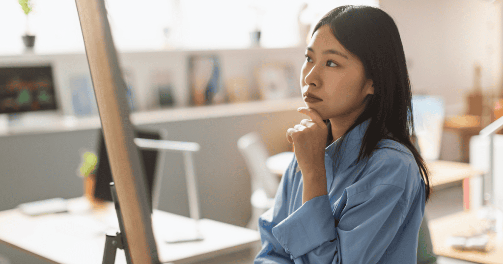 A woman in a blue shirt stands in an office while resting her chin on her hand, appearing to mentally connect dots as she gazes at a board.
