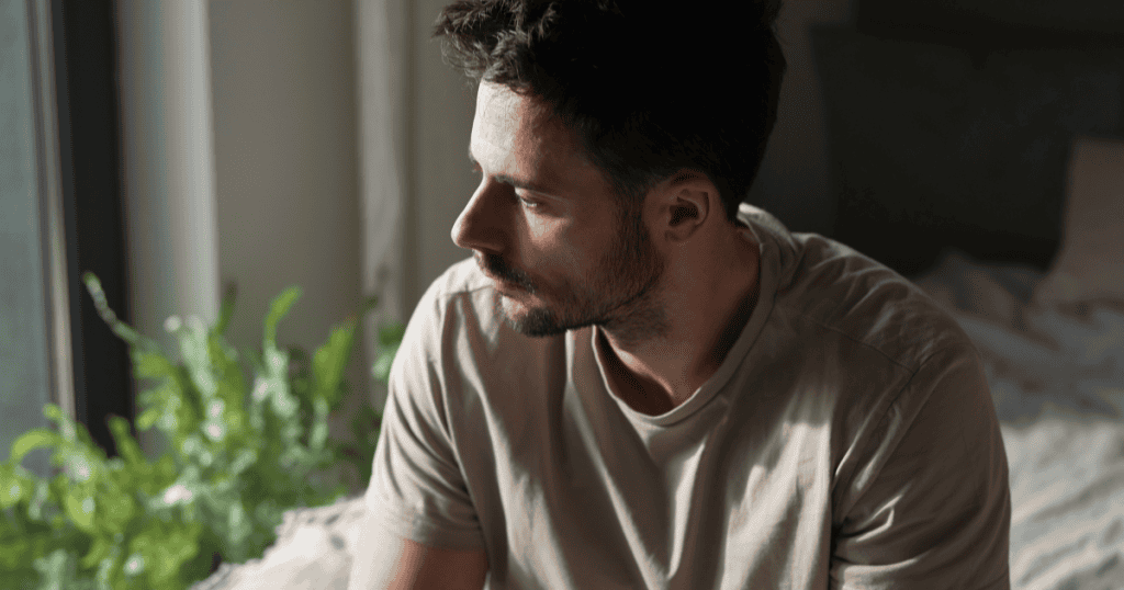A man sits on the edge of a bed and stares out a window into the daylight, mourning the loss of a connection he once took for granted.