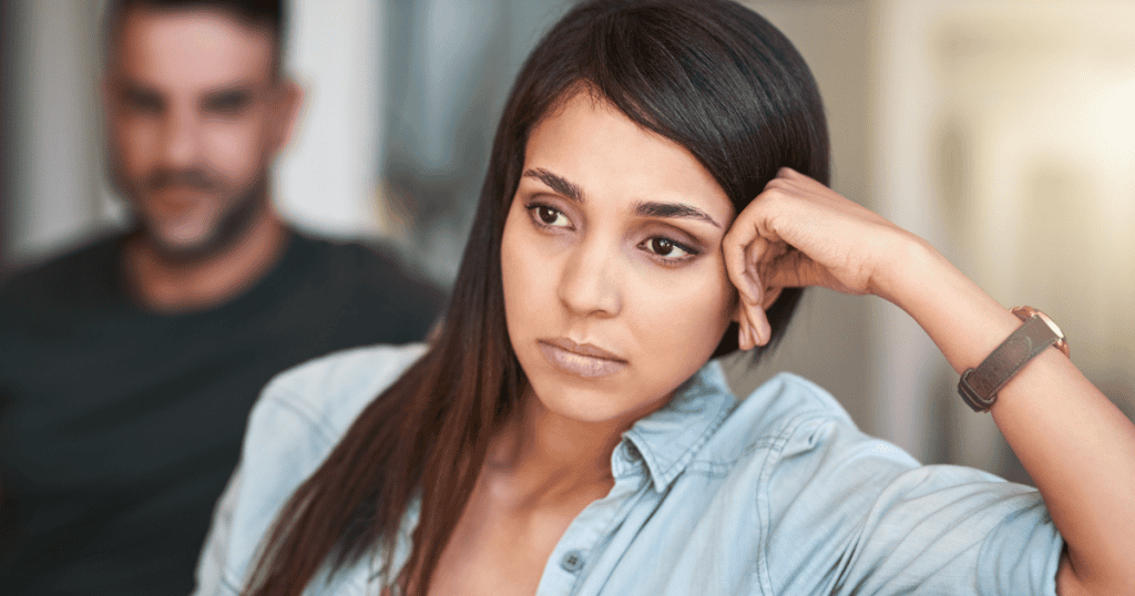A woman gazes off-camera with a weary expression while a man sits blurred in the background, capturing the intuitive weight of an unspoken warning.