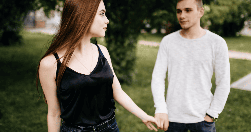 A woman walks away while still holding the hand of a man behind her, portraying a physical disconnect that hasn’t yet reached the fingertips.