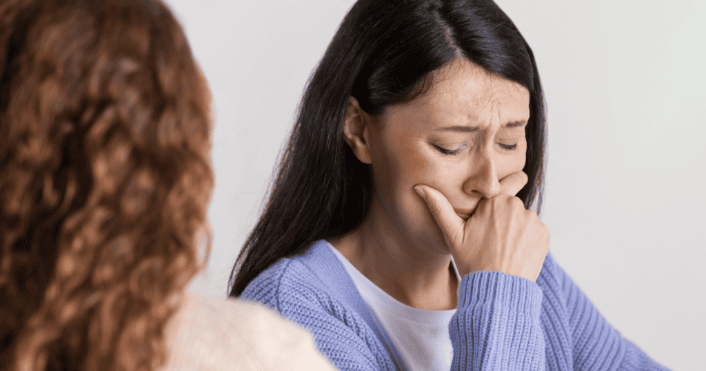 A distressed woman covers her mouth with her hand while listening to someone off-camera, reflecting the quiet strength of stepping back and letting a narcissist unravel on their own.