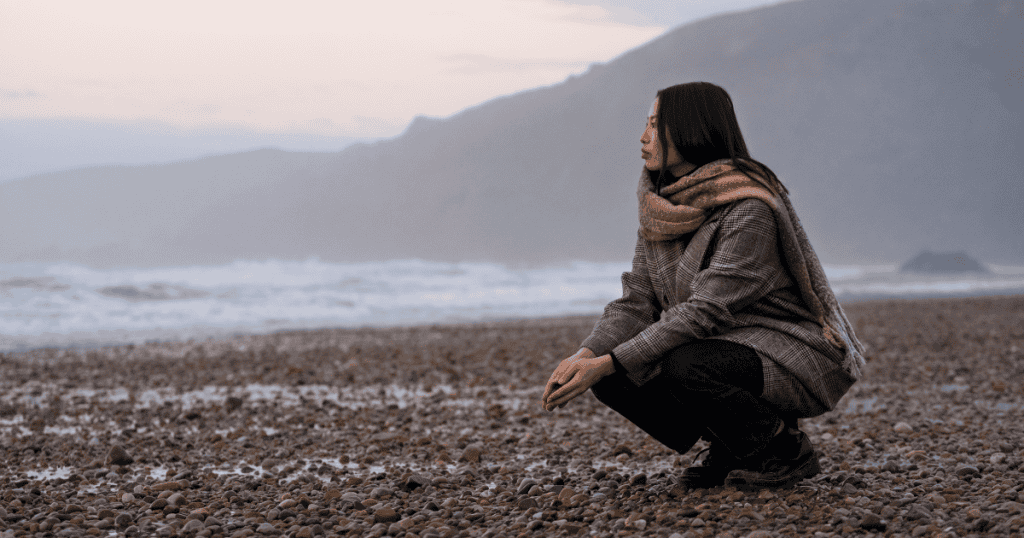 A woman crouches alone on a vast, misty pebble beach looking toward the ocean; the wide-open landscape emphasizes the strength found in solitude.