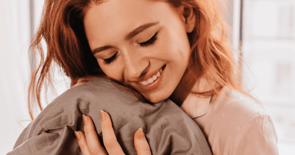 A smiling woman with red hair closes her eyes and hugs a pillow tightly; her peaceful demeanor conveys a sense of internal quiet.