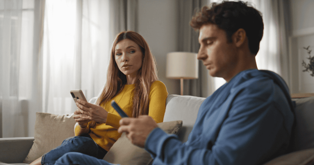 A woman watches a man intensely as he looks at his phone; her cautious gaze suggests a constant, unspoken pressure to anticipate his needs.