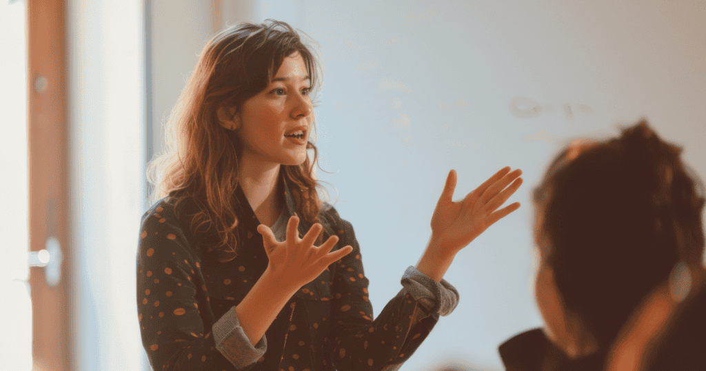 A young woman speaks animatedly with open palms toward an out-of-focus listener, as if she is carefully constructing a logic to justify an uncomfortable reality.