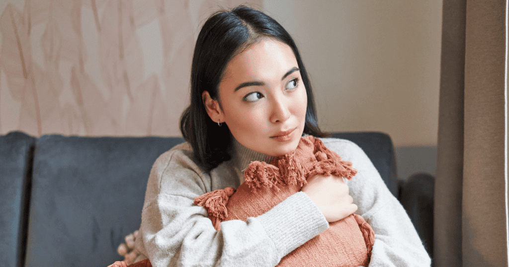 A young woman sits on a sofa hugging a pillow while looking thoughtfully to the side, reflecting on a more measured way to communicate her thoughts.