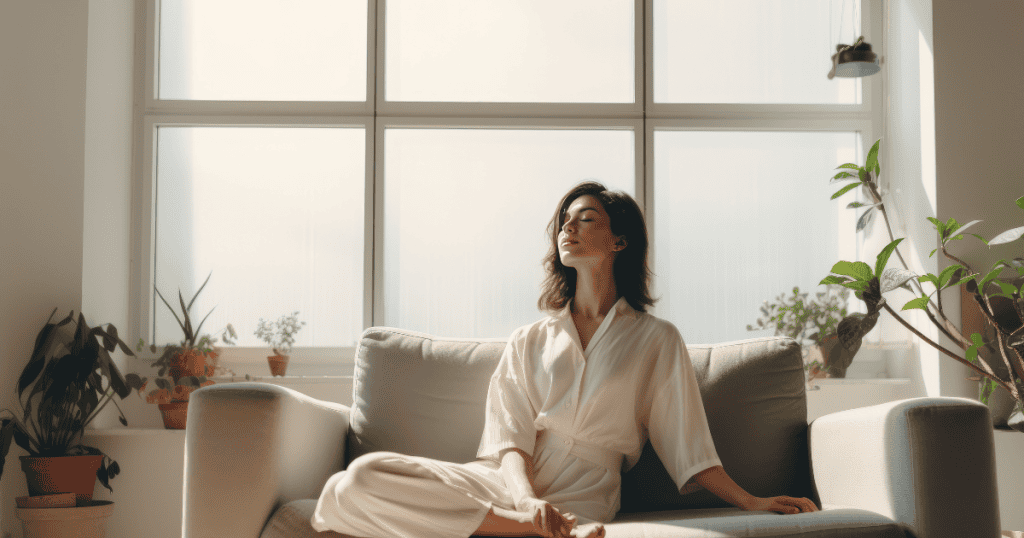 A woman sits peacefully on a sofa with her eyes closed in a sunlit room, her calm posture signaling a quiet restoration of personal sanctuary.