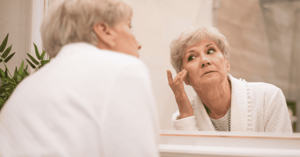 An older woman scrutinizes her reflection in a mirror, her intense gaze reflecting a growing internal friction as time passes.
