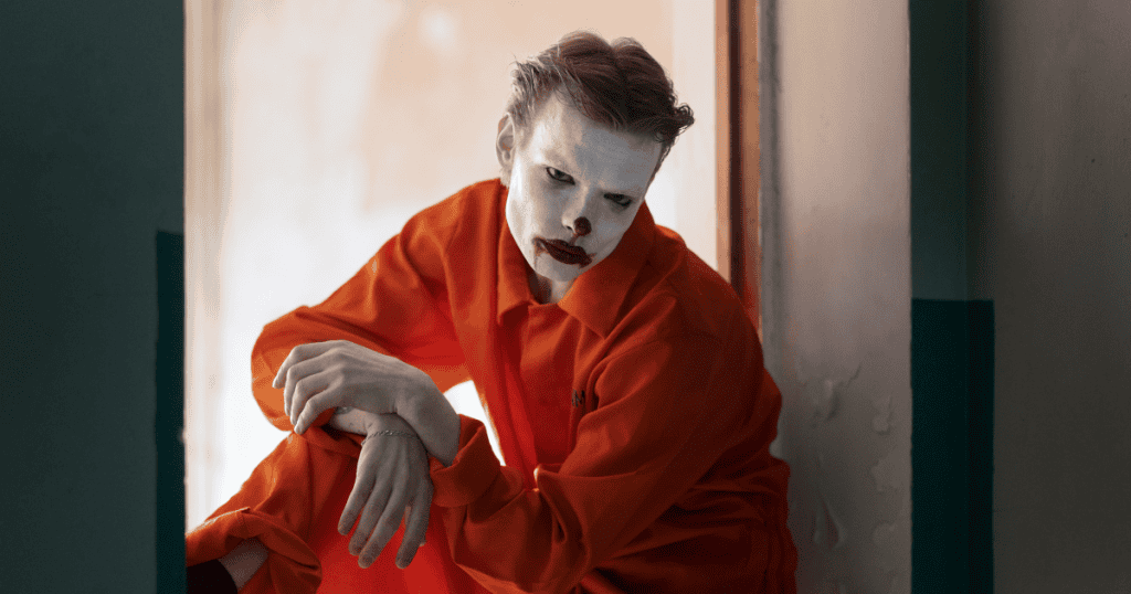A man in an orange jumpsuit wears smeared clown makeup while sitting in a hallway, presenting a theatrical display of suffering that masks a deeper, more calculated intent.