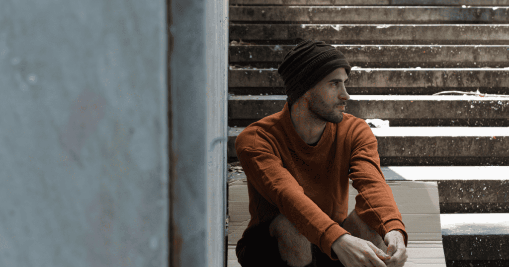 A man in a beanie sits alone on a piece of cardboard on concrete stairs, his quiet posture projecting a sense of perceived victimhood that draws the observer in.
