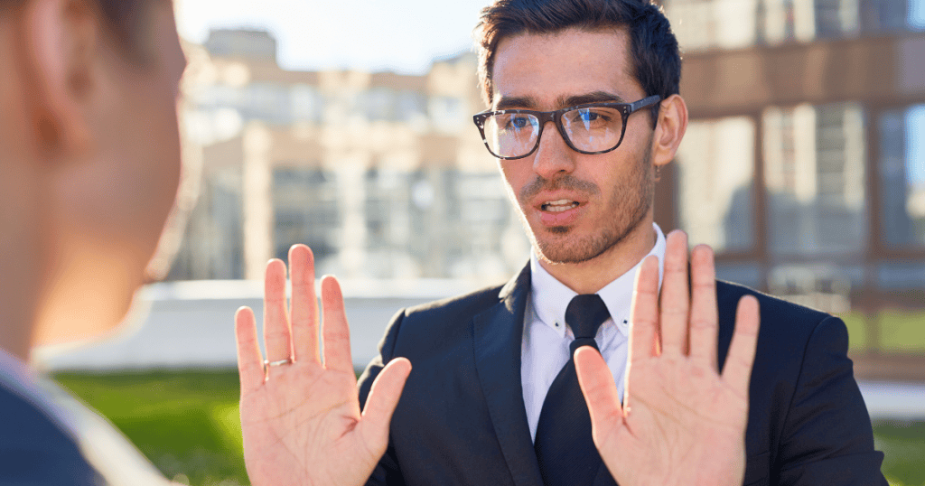 A man in a suit holds his hands up in a defensive "stop" gesture, signaling a rigid refusal to move from his established position.