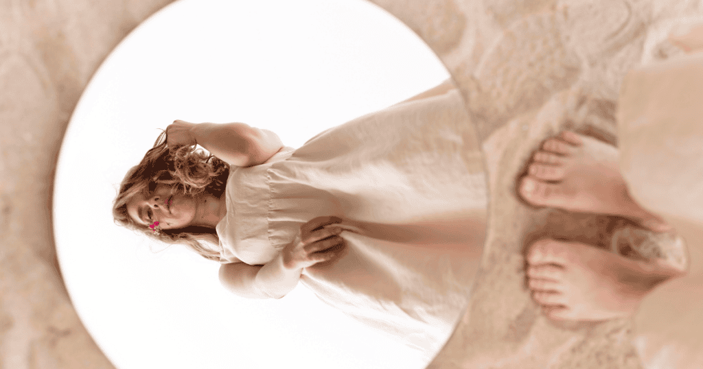 A woman’s reflection is seen in a circular mirror lying on the sand, suggesting the many distorted ways one might see themselves through another's gaze.