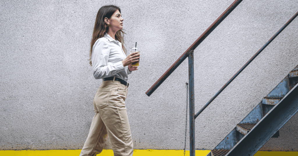 A woman walks purposefully past a set of industrial stairs, her steady pace belying the mounting complexity of leaving a situation behind.