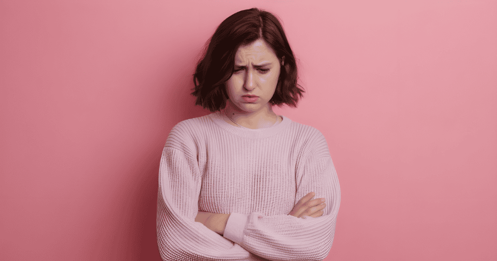 A woman stands against a pink wall with her arms crossed and a look of deep shame, portraying the heavy discomfort that can come from simply trying to be happy.