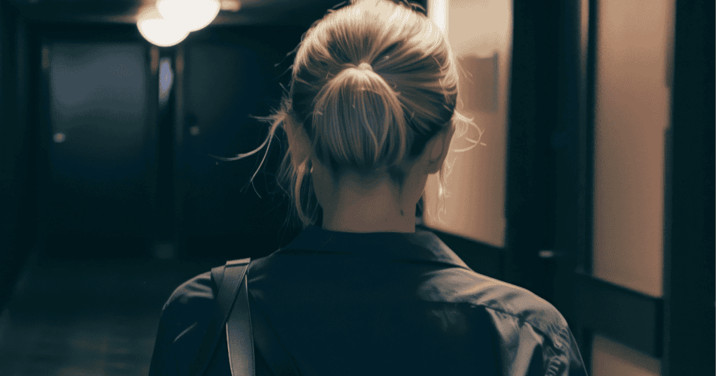 A view from behind shows a woman standing at the end of a long, shadowy hallway, illustrating the daunting psychological distance between staying and taking that first step away.