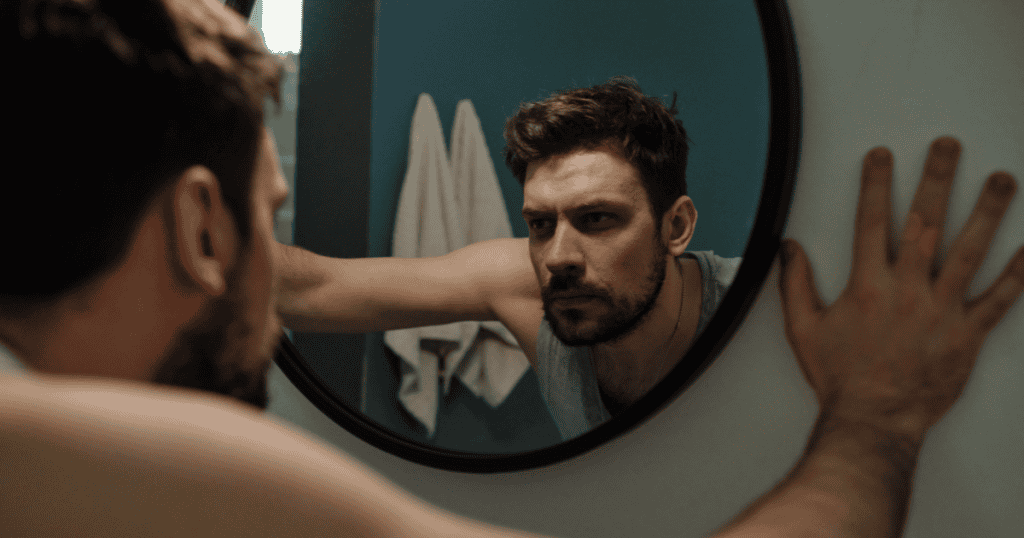 A man leans heavily against a bathroom mirror while staring at his reflection, confirming a grim reality that has finally come into focus.