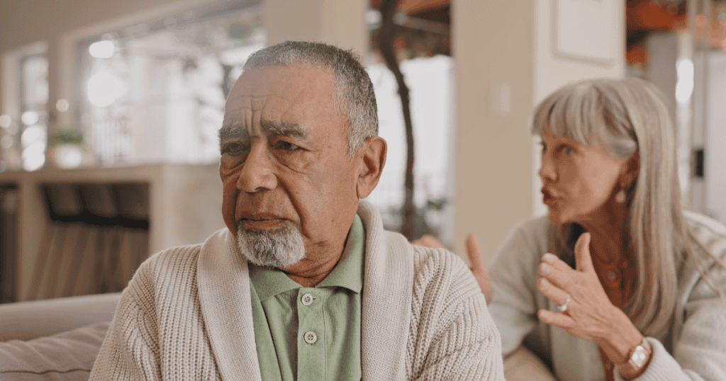 An older man stares blankly into the distance while a woman gestures emphatically behind him, reflecting a quiet withdrawal as a once-dominant presence turns into cold resentment.
