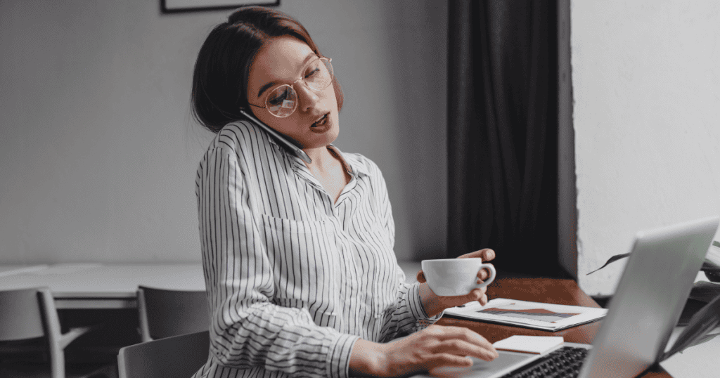 A woman in a striped shirt juggles a phone call and a laptop while holding a coffee cup, her strained expression suggesting the hidden weight of maintaining an impossible balance.