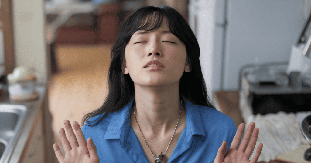 A young woman stands in a kitchen with her eyes closed and palms facing outward, taking a grounding breath to maintain her internal center against the surrounding environment.