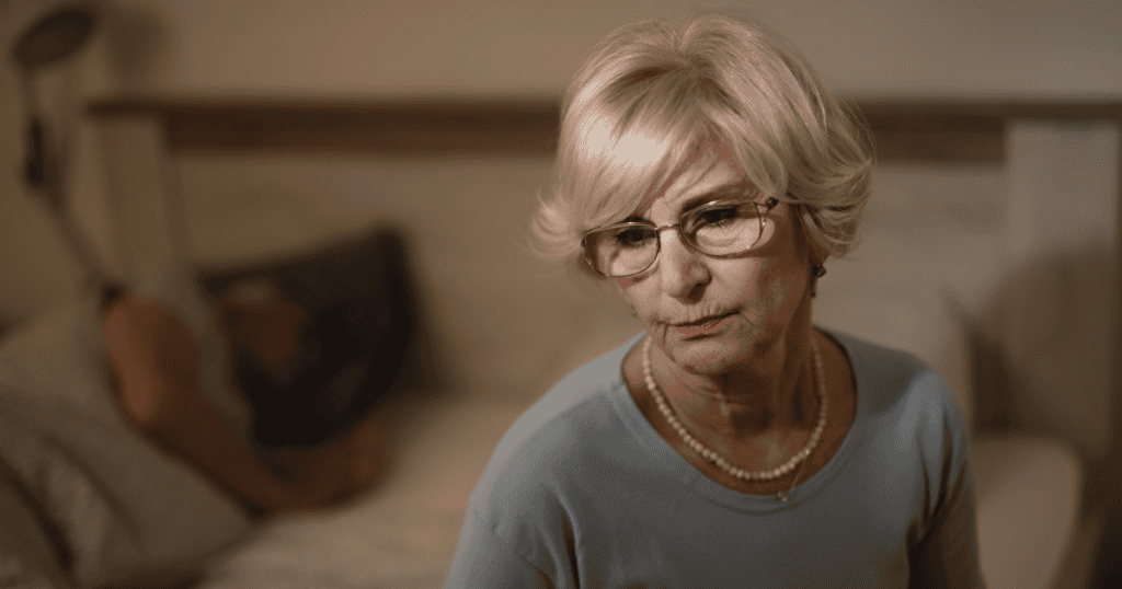 An older woman sits in silence while someone rests in the background, reflecting the emotional heaviness and isolation that can deepen with aging narcissistic behavior.