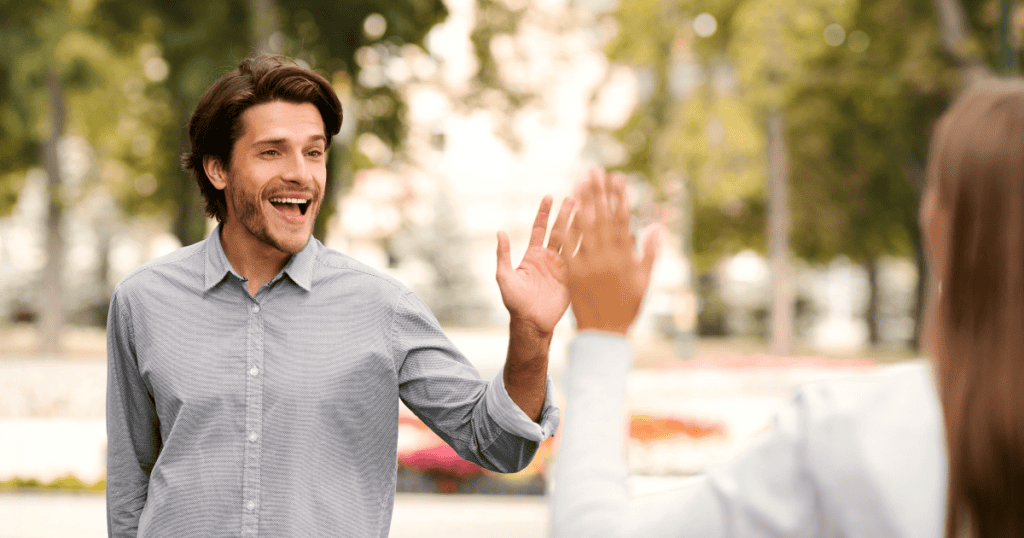 A man smiles broadly and raises his hand in a high-five gesture toward a woman whose back is to the camera, illustrating the disarming and confusing nature of a sudden, cheerful reappearance.