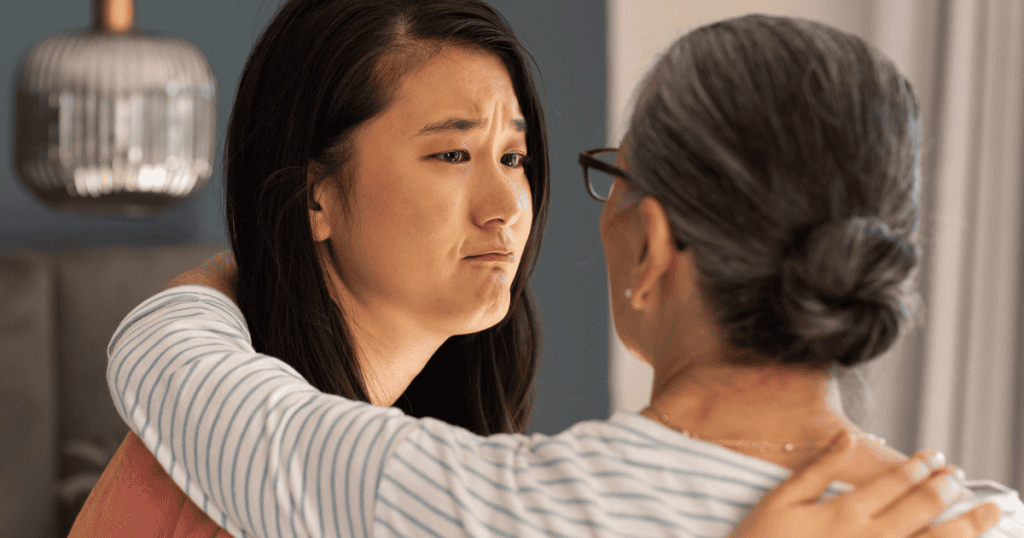 A young woman with a tearful, distressed expression looks down while an older woman wraps an arm around her shoulder, illustrating an early habit of absorbing another person's emotional weight.