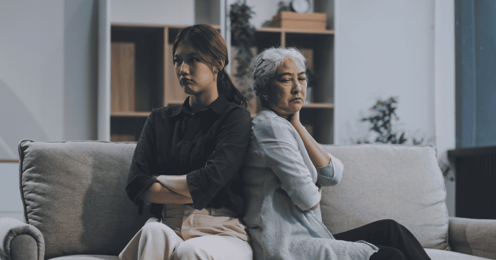 Two women of different generations sit back-to-back on a gray sofa with crossed arms and somber expressions, illustrating the stony silence of a sudden emotional severance.