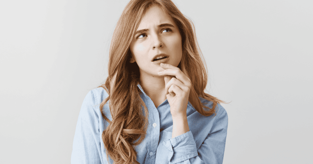A young woman in a blue shirt looks upward with a skeptical, pondering expression and her finger on her chin, illustrating the deep contemplation that happens right before deciding whether to buy into a familiar narrative again.
