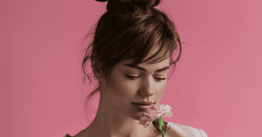 A young woman with her hair in a bun closes her eyes and gently smells a pink carnation, representing the quiet return to personal pleasure and sensory awareness.