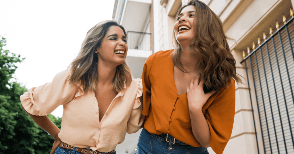 Two young women walk side-by-side down a city street laughing together in genuine joy, capturing the lighthearted origin of an emotional bond before it becomes one-sided.