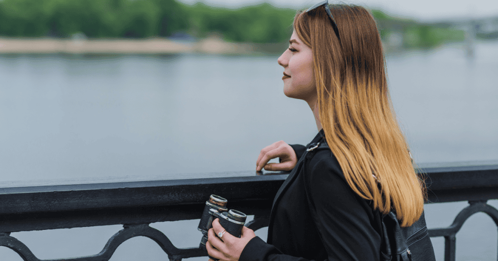 A woman stands at a waterfront railing holding a pair of vintage binoculars and looking out across the river, capturing the wistful act of looking back at the past while remaining anchored on the safe side of the boundary.