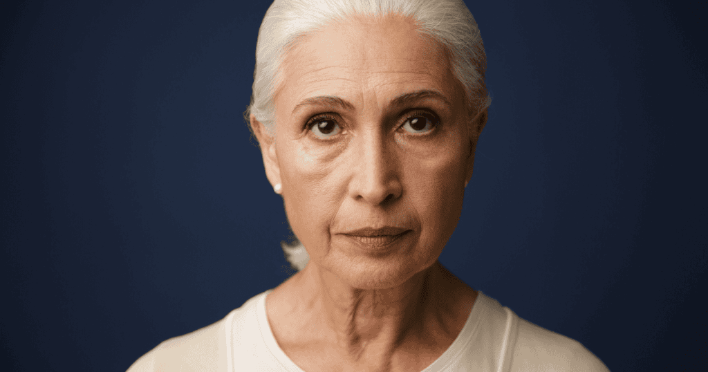 An older woman with pulled-back white hair looks directly forward with an unblinking, neutral gaze, projecting a calculated emotional barrier that forces others to look for approval.
